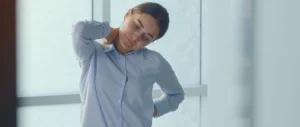 Woman in blue shirt stretching neck at office window, experiencing office syndrome pain