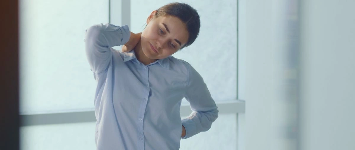 Woman in blue shirt stretching neck at office window, experiencing office syndrome pain